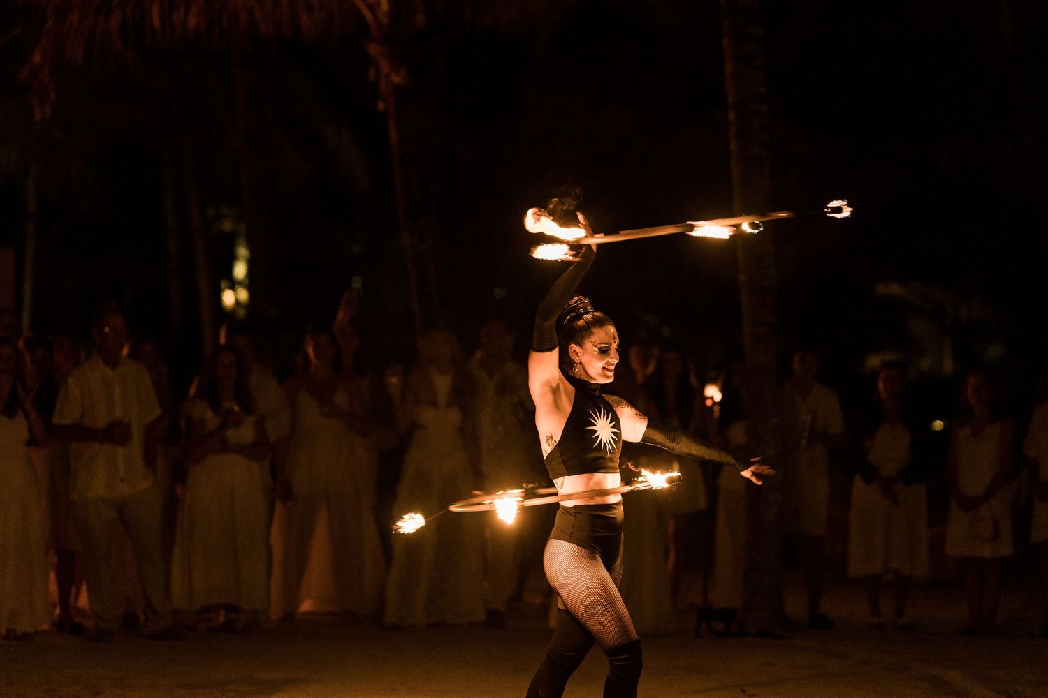 Fire dancer performing at night with flaming batons, surrounded by audience in tropical outdoor setting.