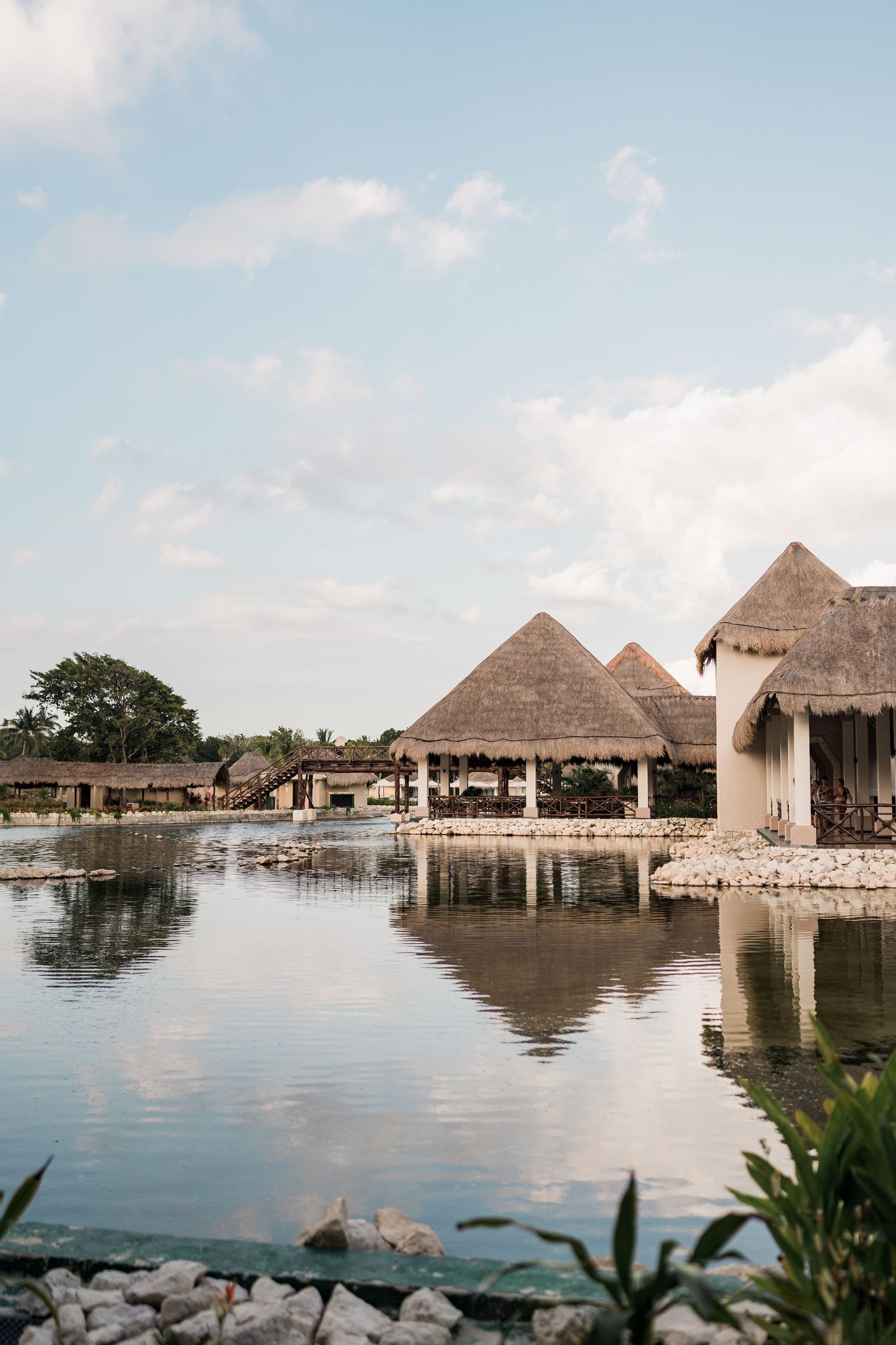 Thatched-roof bungalows reflecting on calm lagoon waters at a tropical resort under a partly cloudy sky.