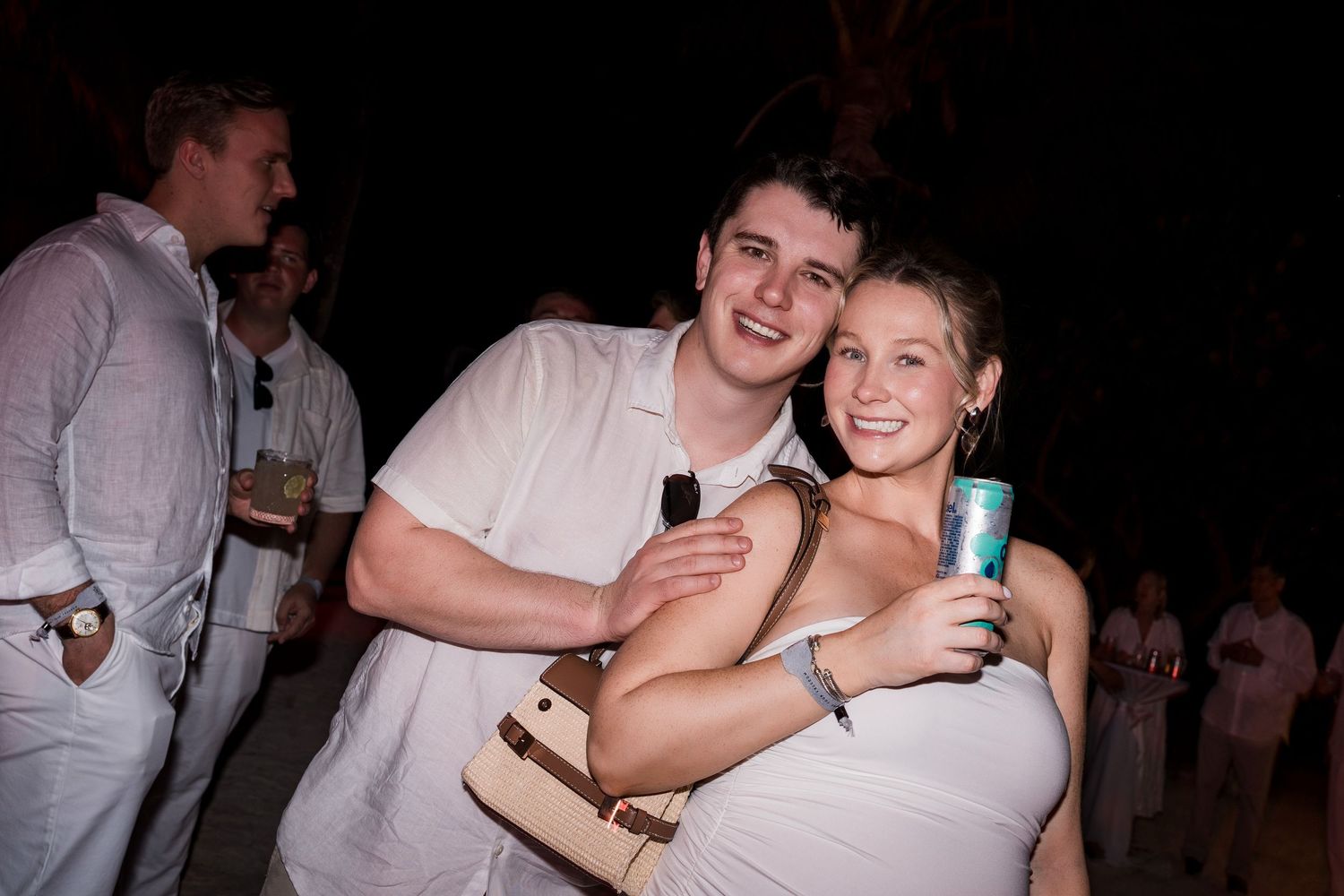 Two smiling young adults in white outfits pose together at an outdoor evening party event.