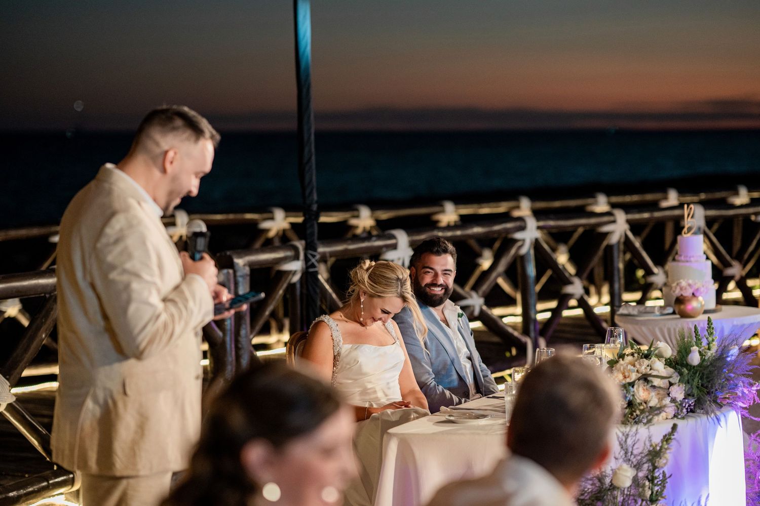 Man giving wedding toast to smiling bride and groom at outdoor reception with ocean sunset backdrop.