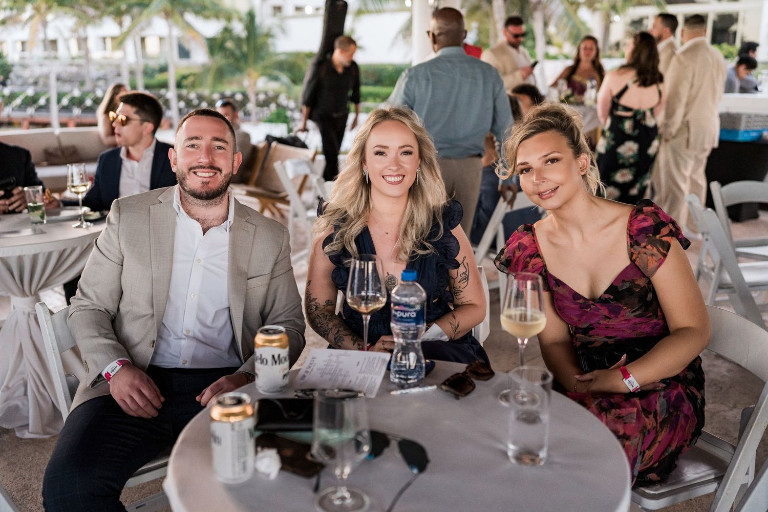 Three guests smile at an outdoor event table, holding champagne glasses at an elegant waterfront venue.