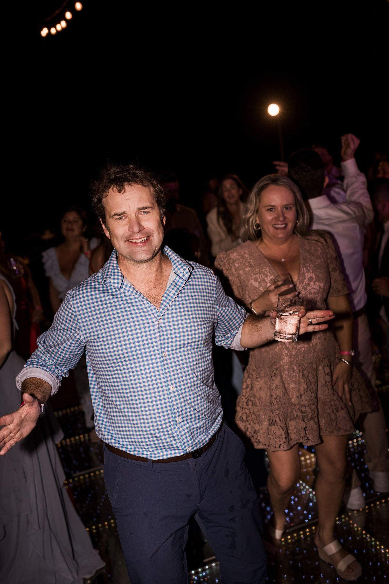 A smiling man in a blue gingham shirt dances at an outdoor evening party with guests celebrating behind him.