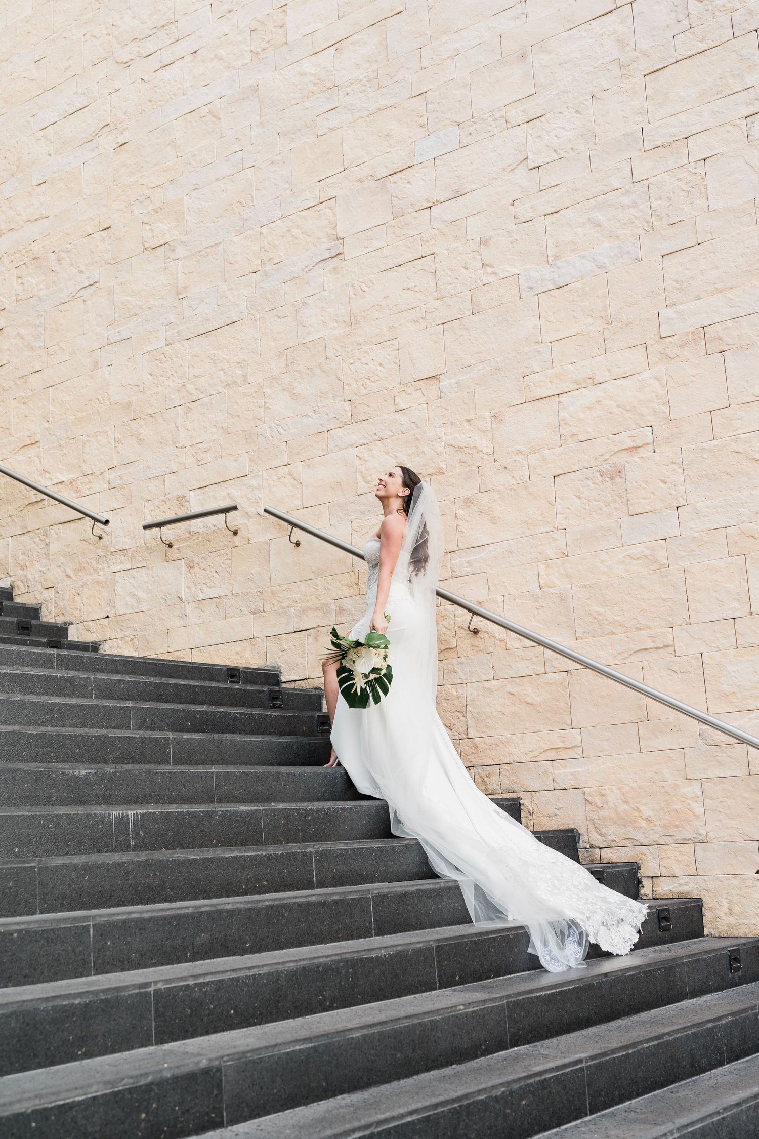 Bride in white gown holding bouquet ascending stone steps against limestone wall.