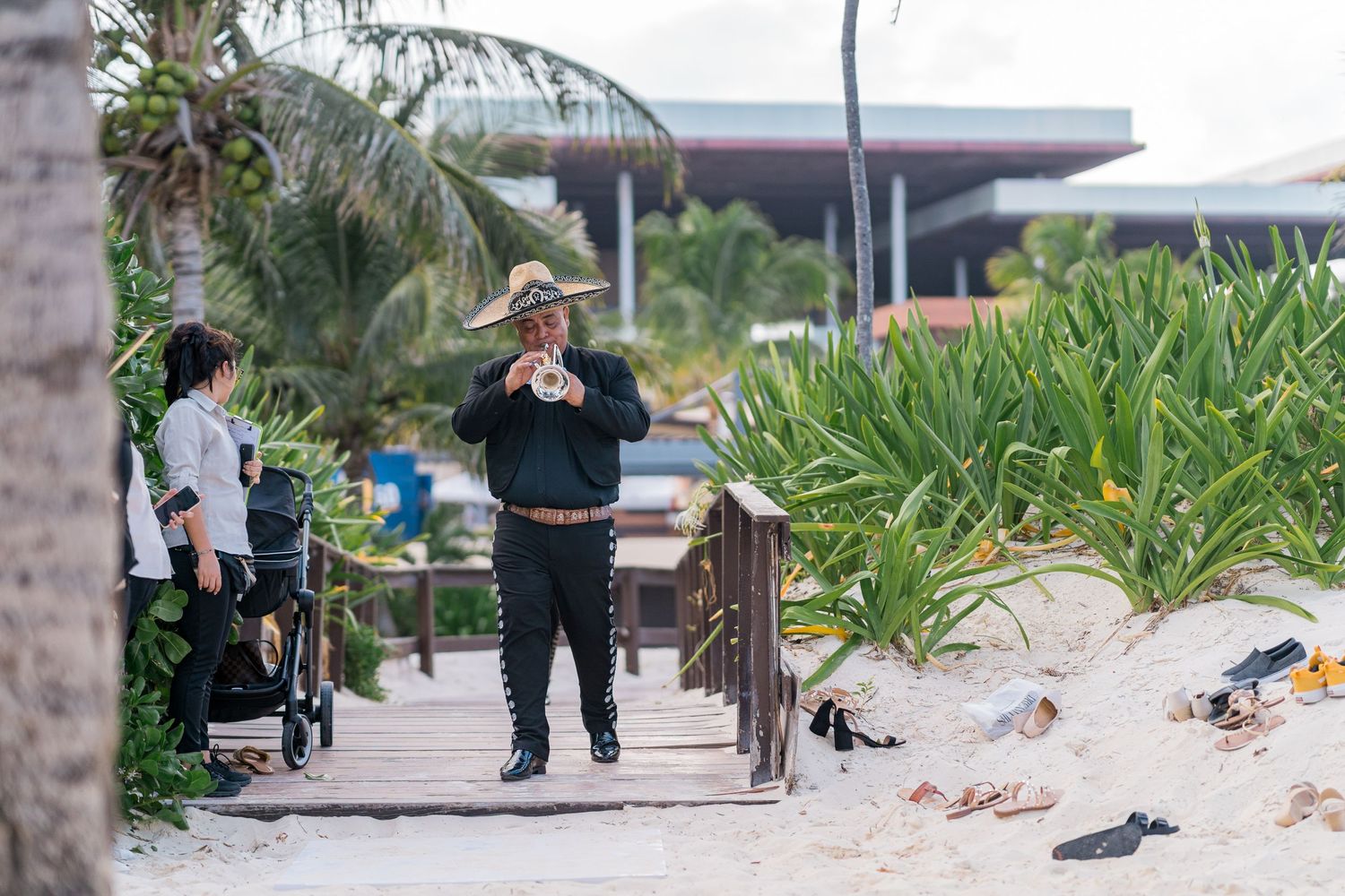 Musician in sombrero plays instrument on a sandy beachside boardwalk surrounded by tropical palm trees.