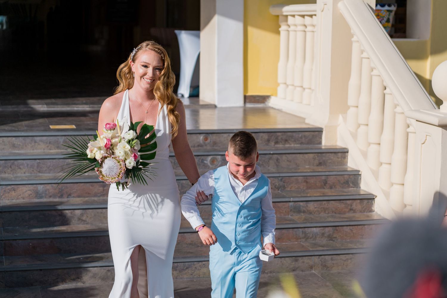Smiling blonde bride in a white gown walking down steps with a young boy in a light blue suit.