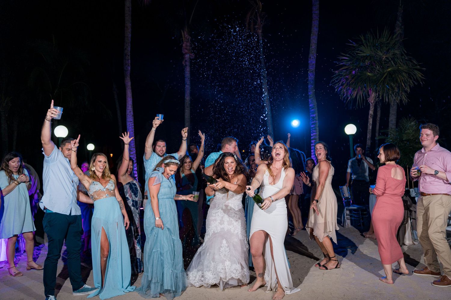 Bride and wedding guests dancing joyfully on sandy beach at nighttime outdoor reception.