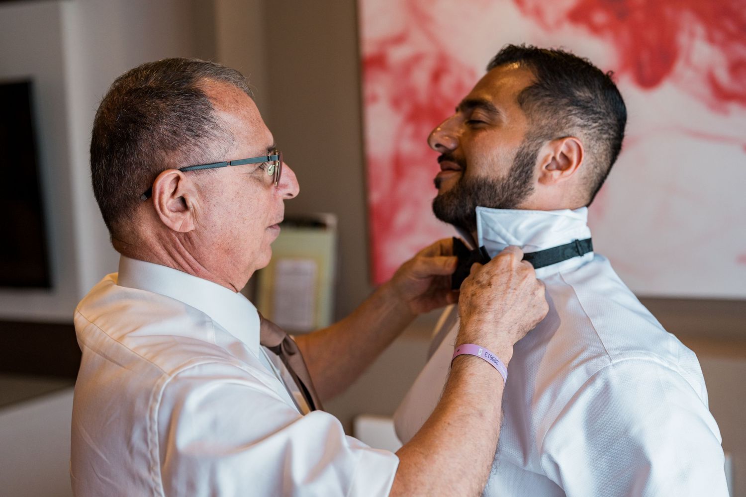 Father helps groom adjust bow tie during wedding day preparations indoors.