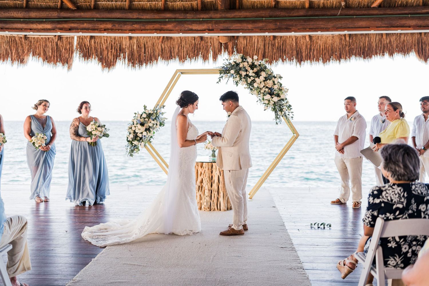 Bride and groom exchange vows at hexagonal floral arch with ocean backdrop.