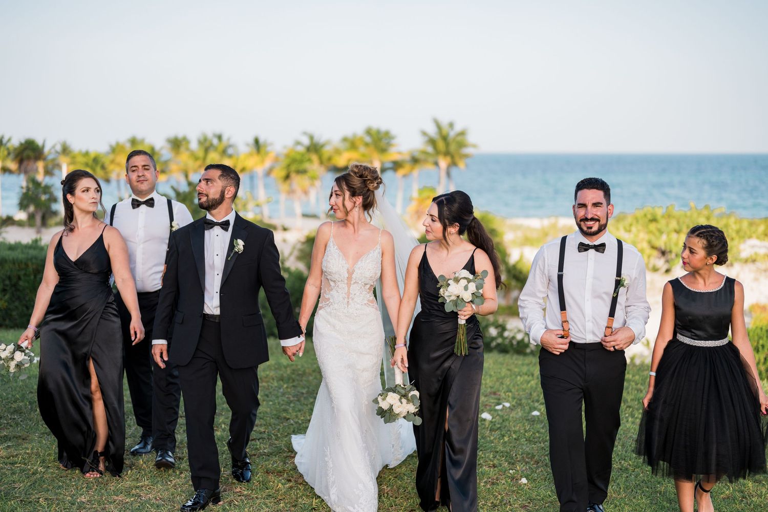 Wedding party walks together on lush green lawn with tropical ocean scenery in the background.