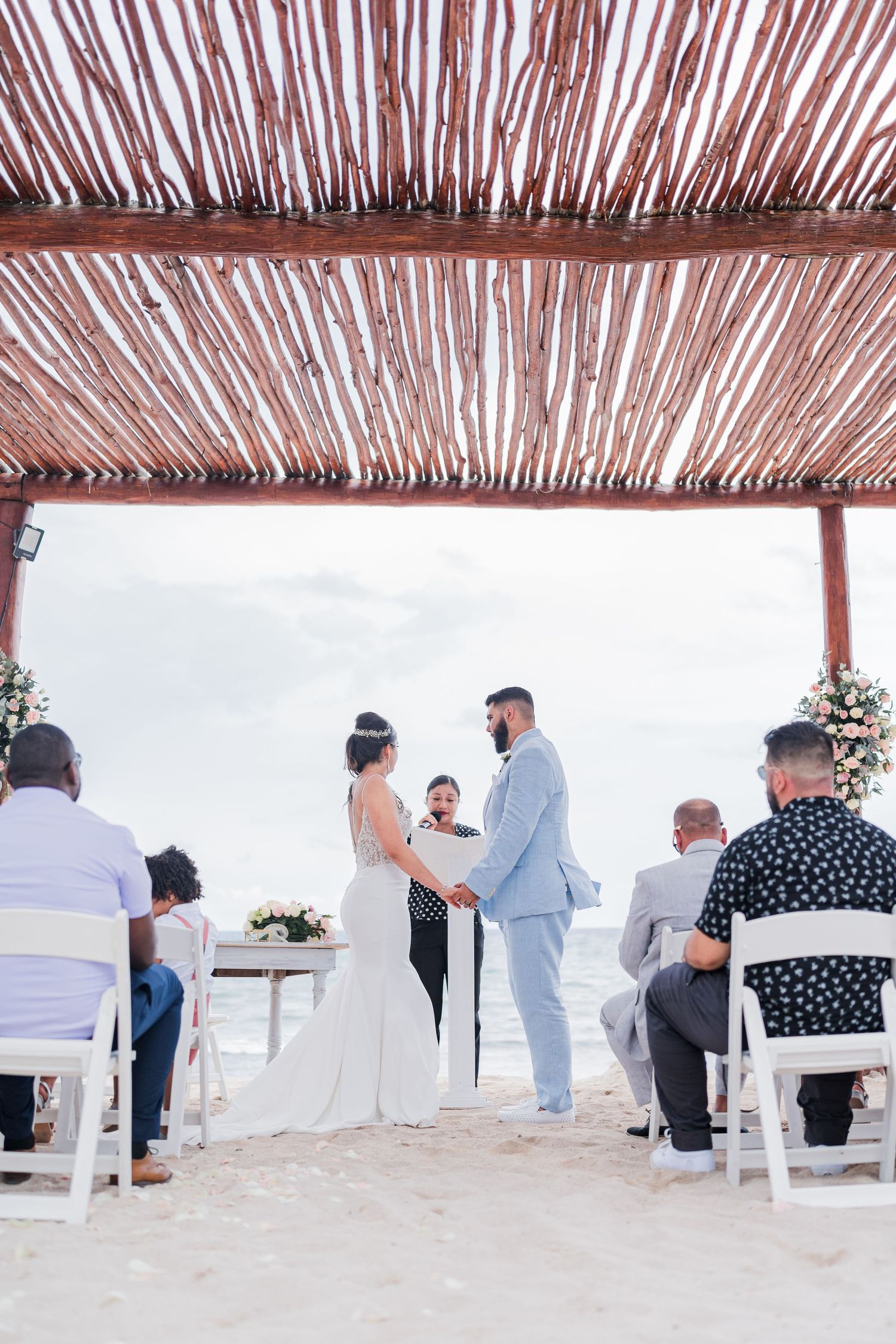 Couple holds hands during beach wedding ceremony under rustic wooden pergola with ocean view backdrop.