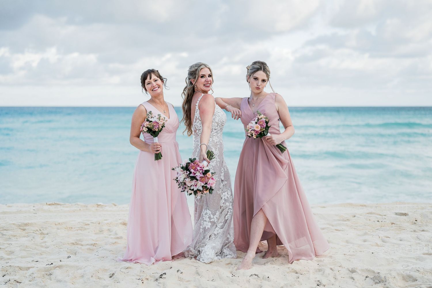 Bride in lace gown posing with two bridesmaids in blush pink dresses on sandy beach.