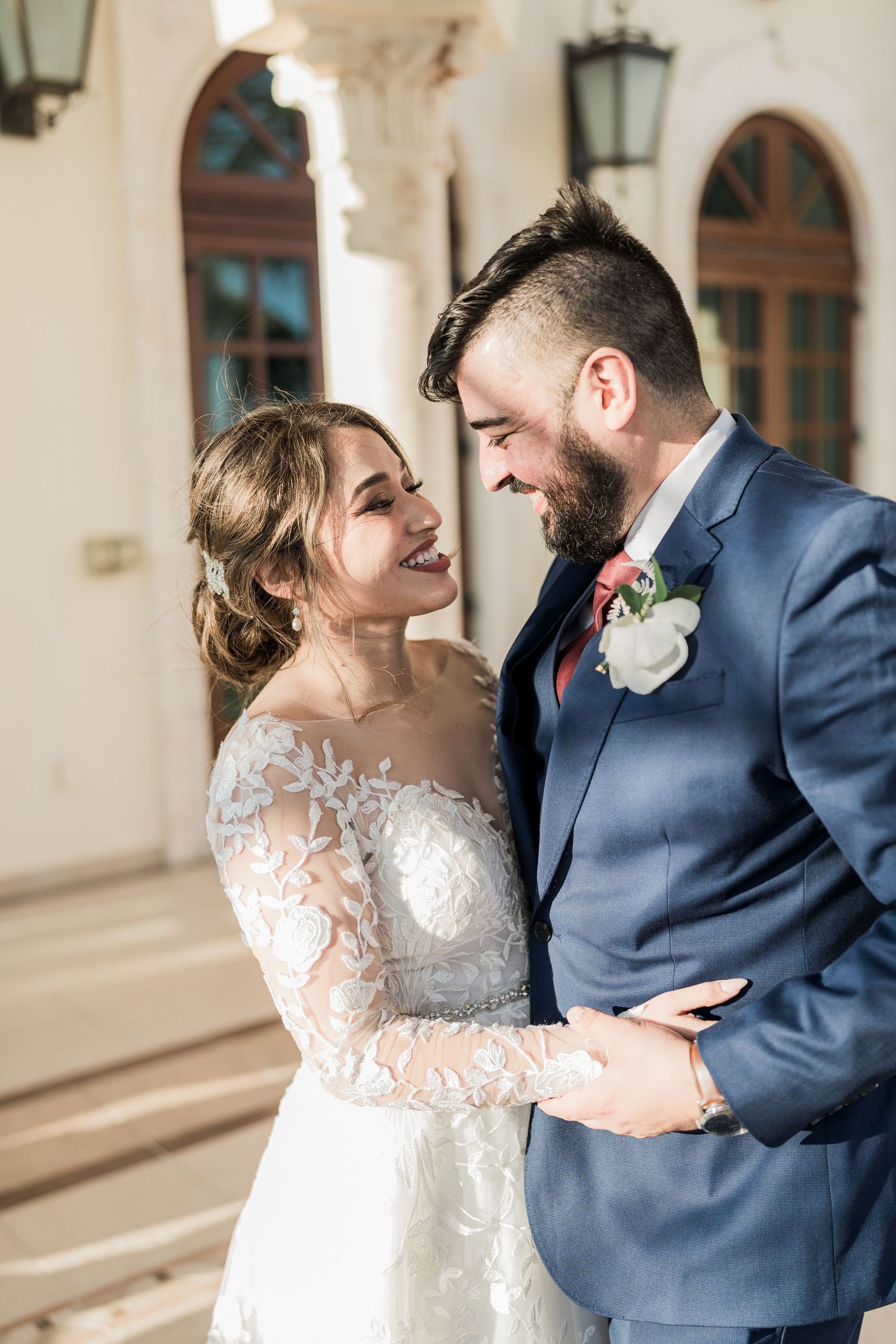 Laughing bride in lace gown and bearded groom in navy suit sharing joyful moment at wedding venue.