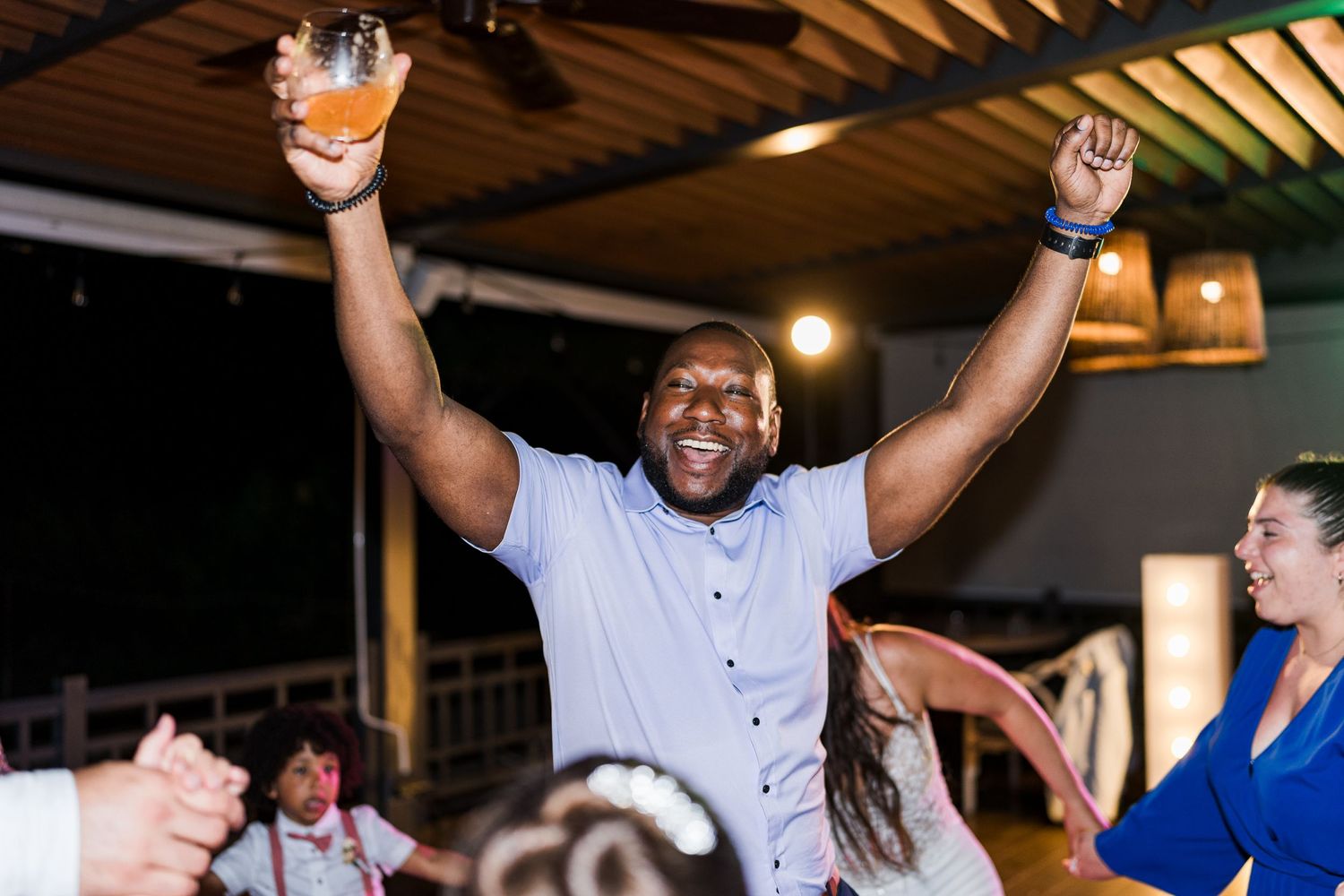 Joyful wedding guest raises arms and drink in celebration on the dance floor.