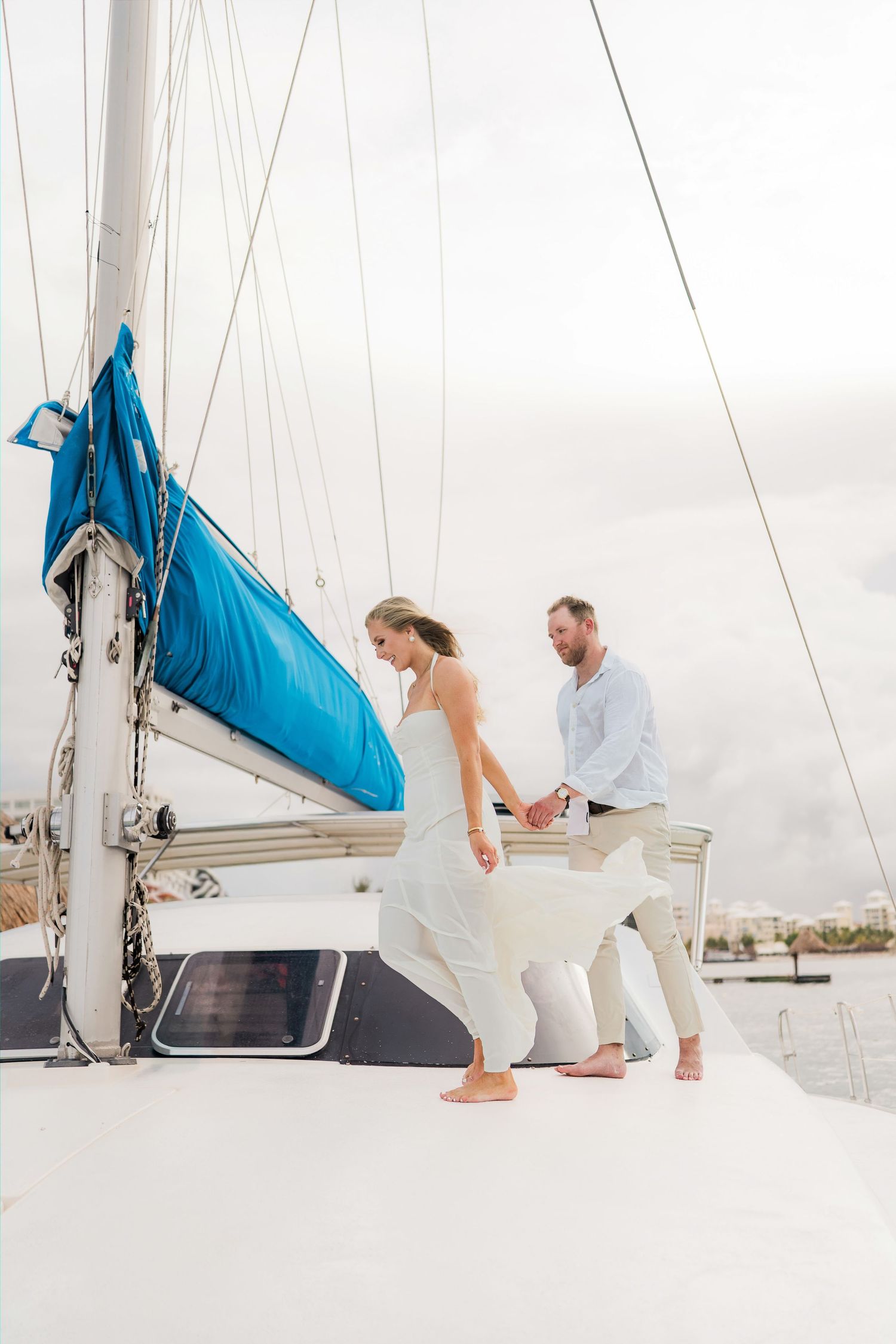 Cancun Engagement Photoshoot - Barefoot bride and groom holding hands on a white sailboat deck with blue sails on a cloudy day.