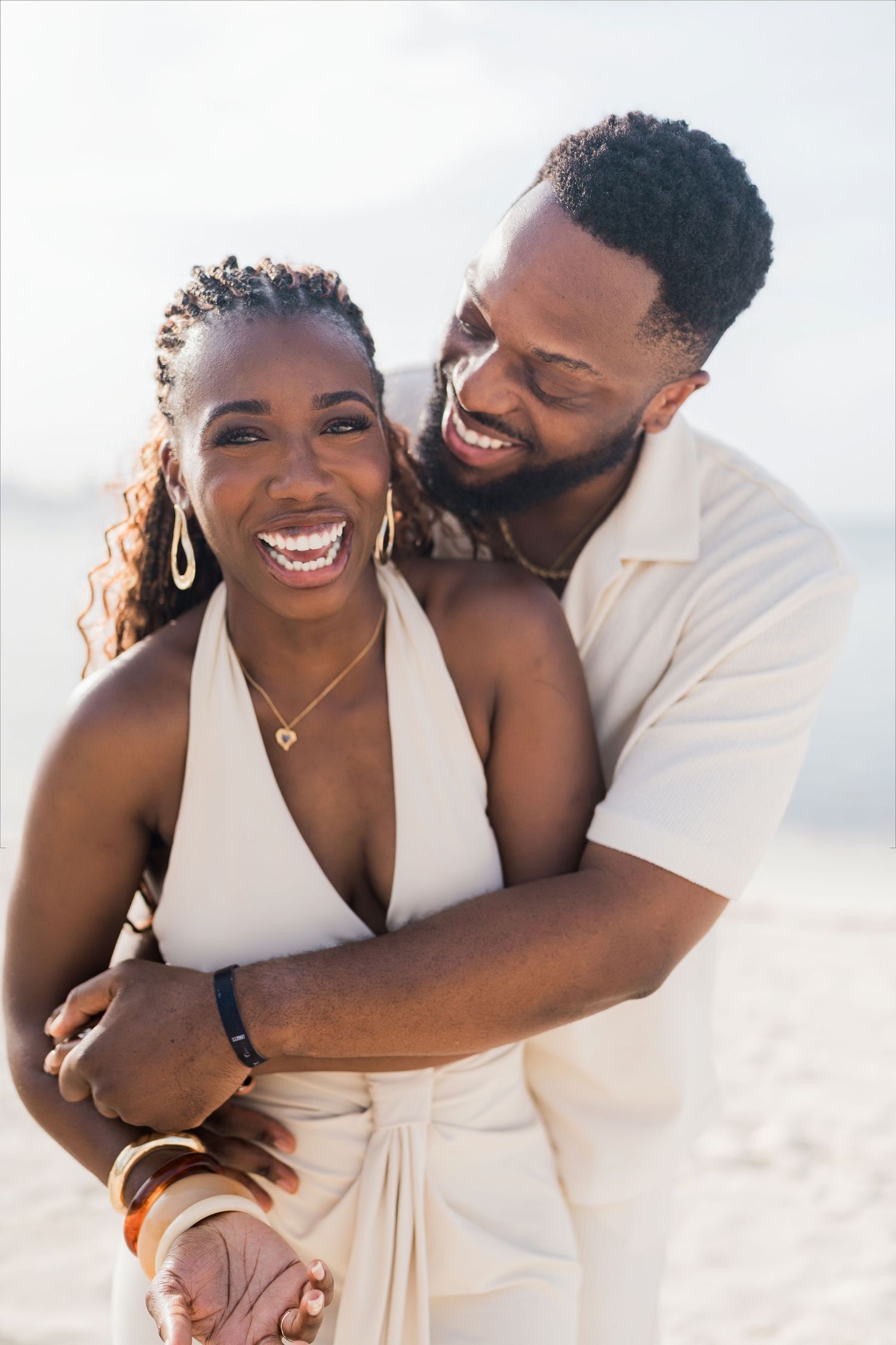 A joyful Black couple embraces and laughs together on a bright sandy beach.