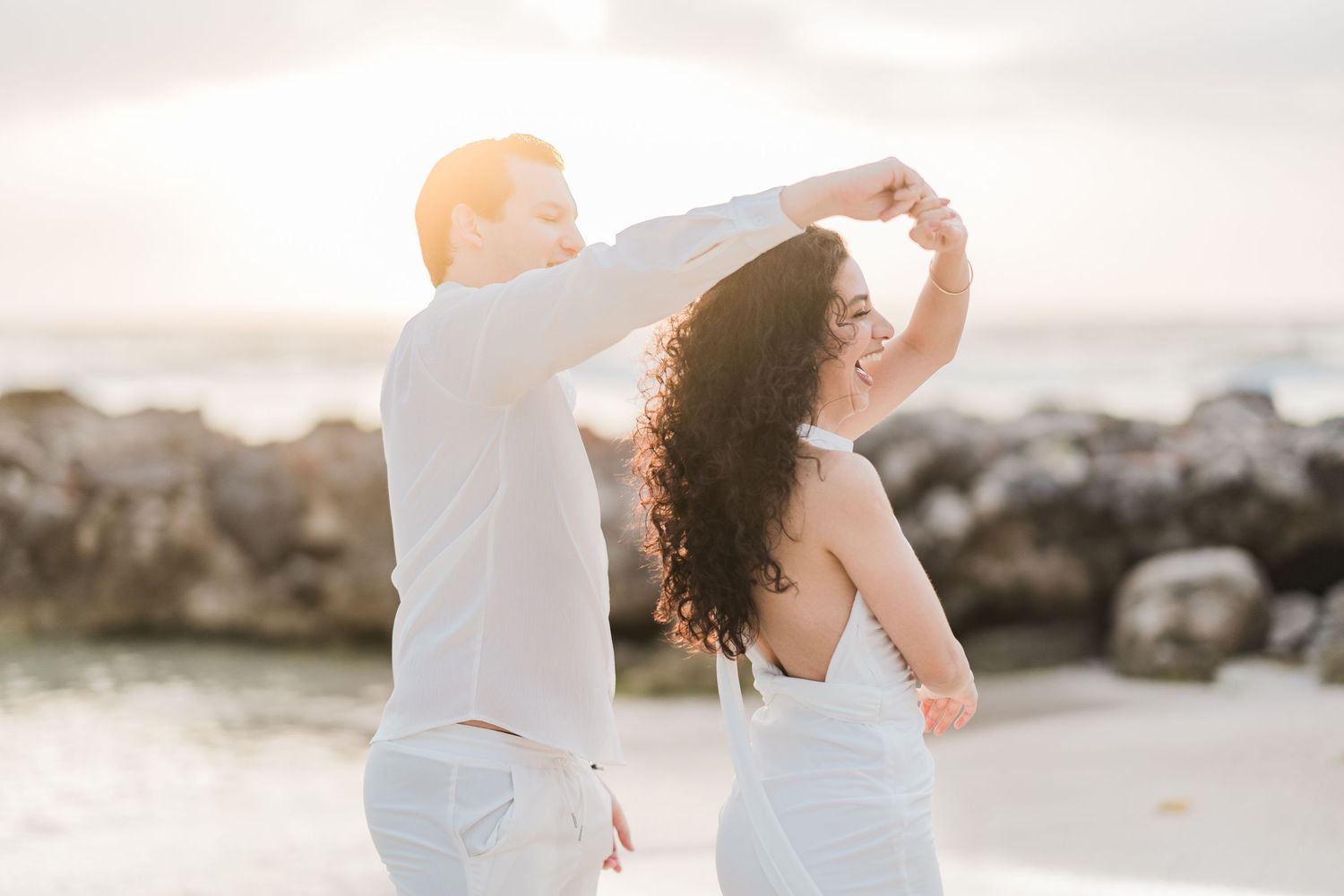 Couple dancing on a beach at sunset, woman in white dress being twirled by man in white outfit.