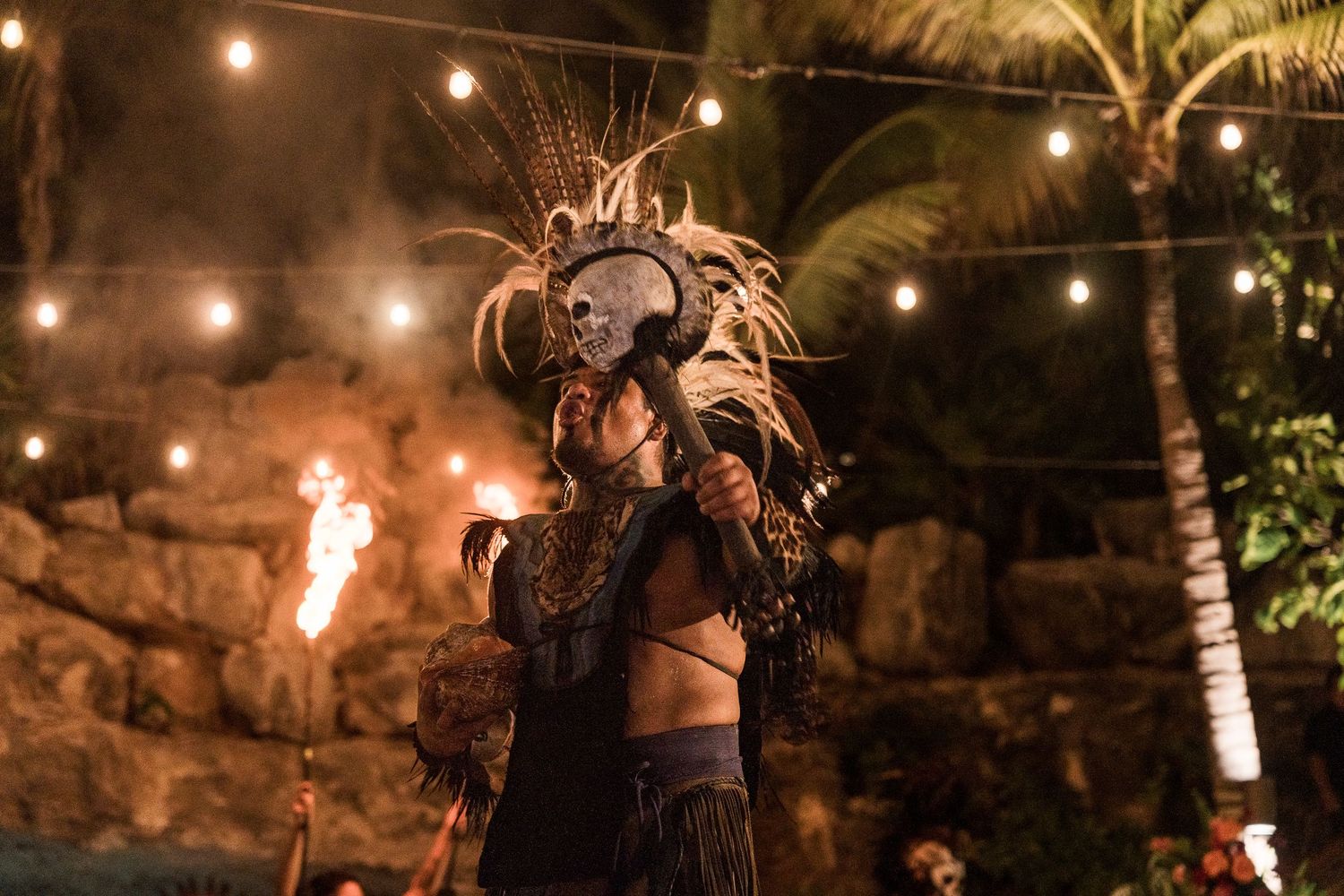 Performer in elaborate Mayan warrior costume with skull mask and feathered headdress at a nighttime cultural show.