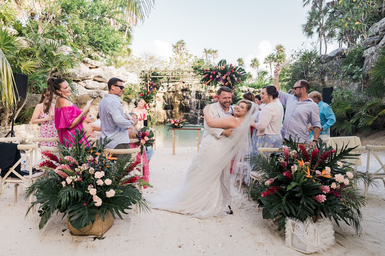 Couple shares first kiss at tropical beach wedding ceremony with lush floral arrangements and guests seated outdoors.
