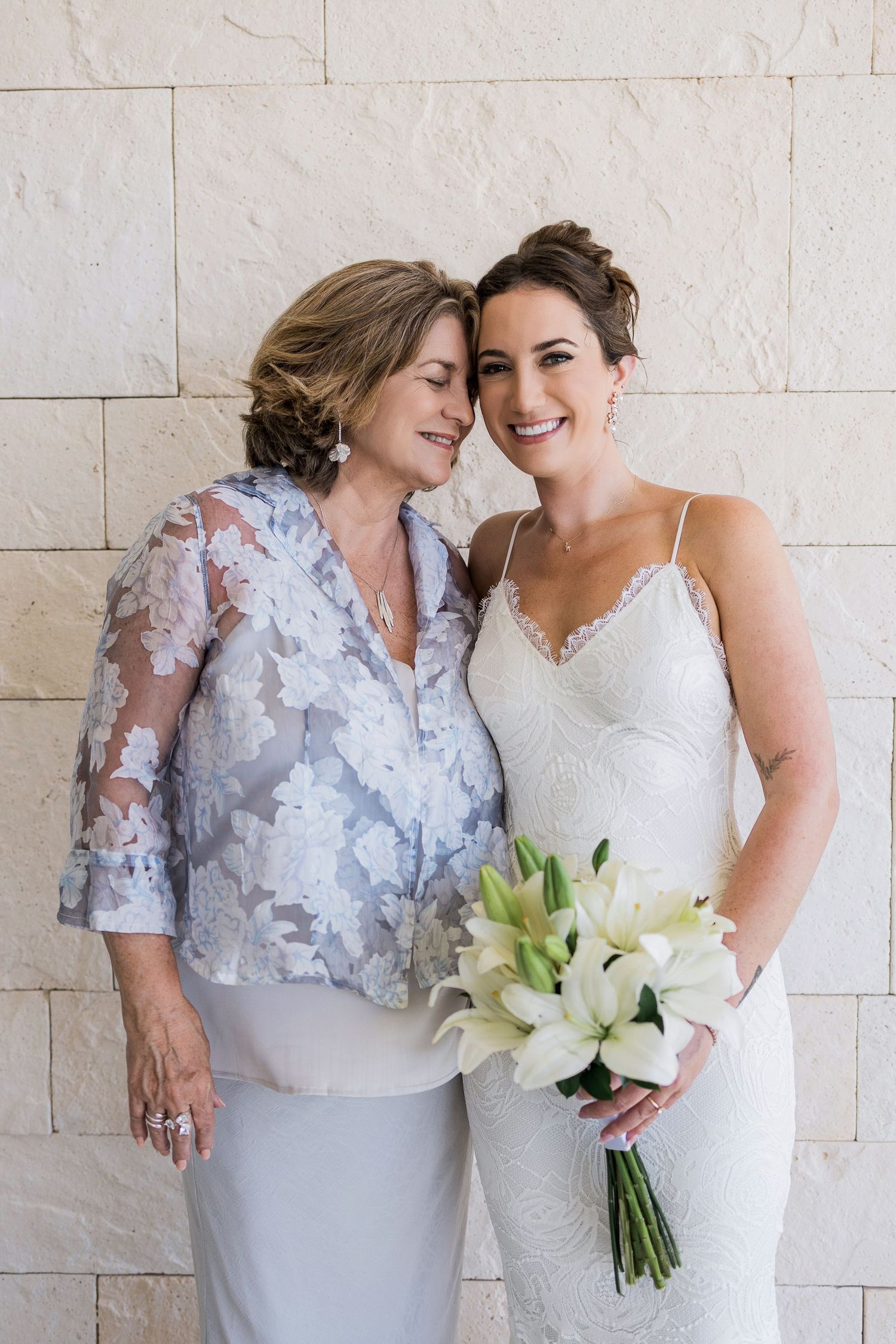 Bride in white lace gown holding white lily bouquet poses with older woman in blue floral jacket against stone wall.
