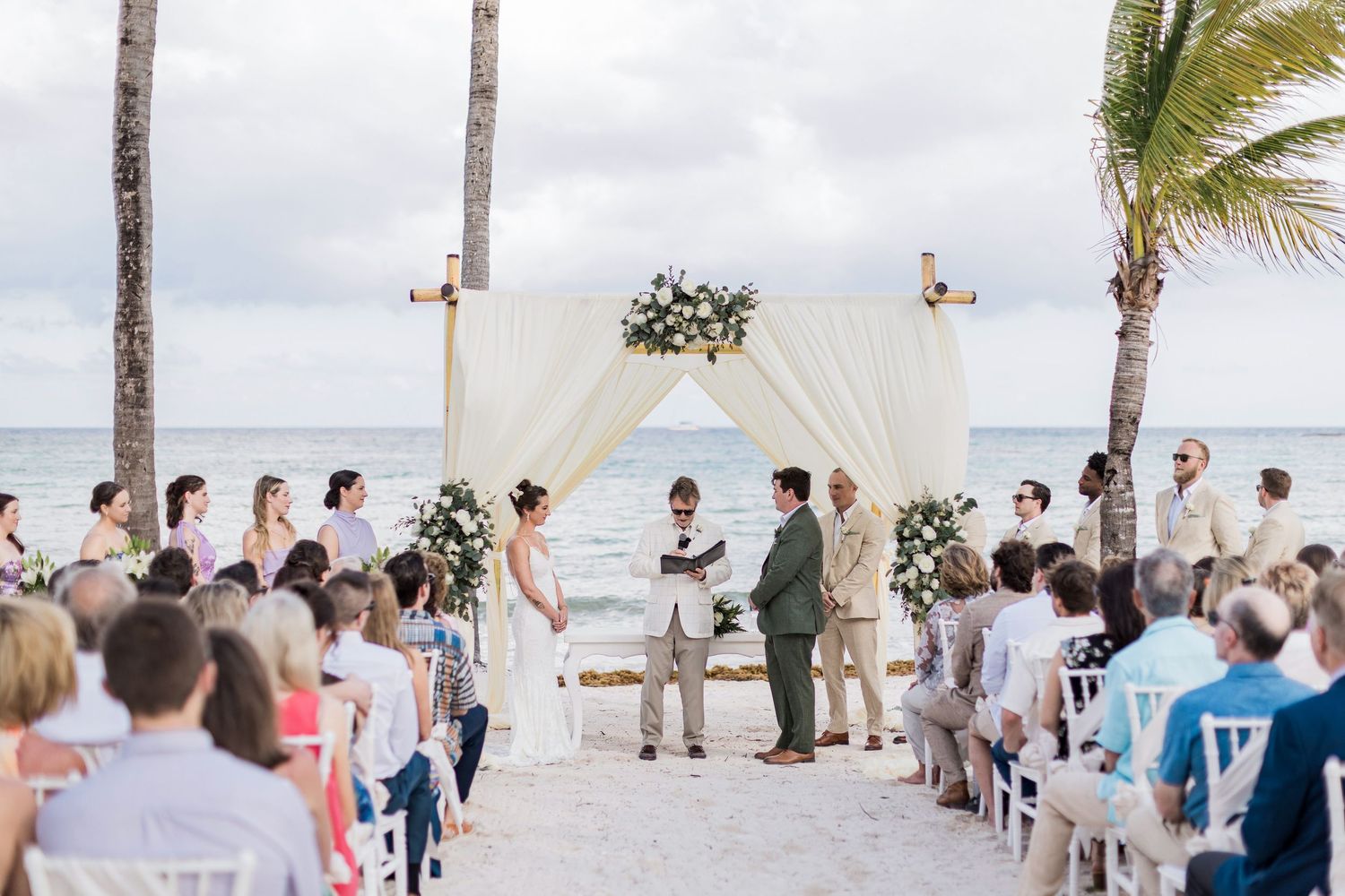 Romantic beach wedding ceremony with couple at floral arch, guests seated on white chairs, ocean backdrop, and palm trees.