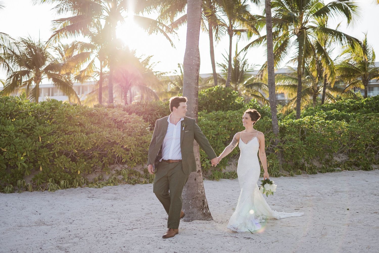 Bride and groom hold hands walking on a tropical beach wedding with palm trees and golden sunset light.