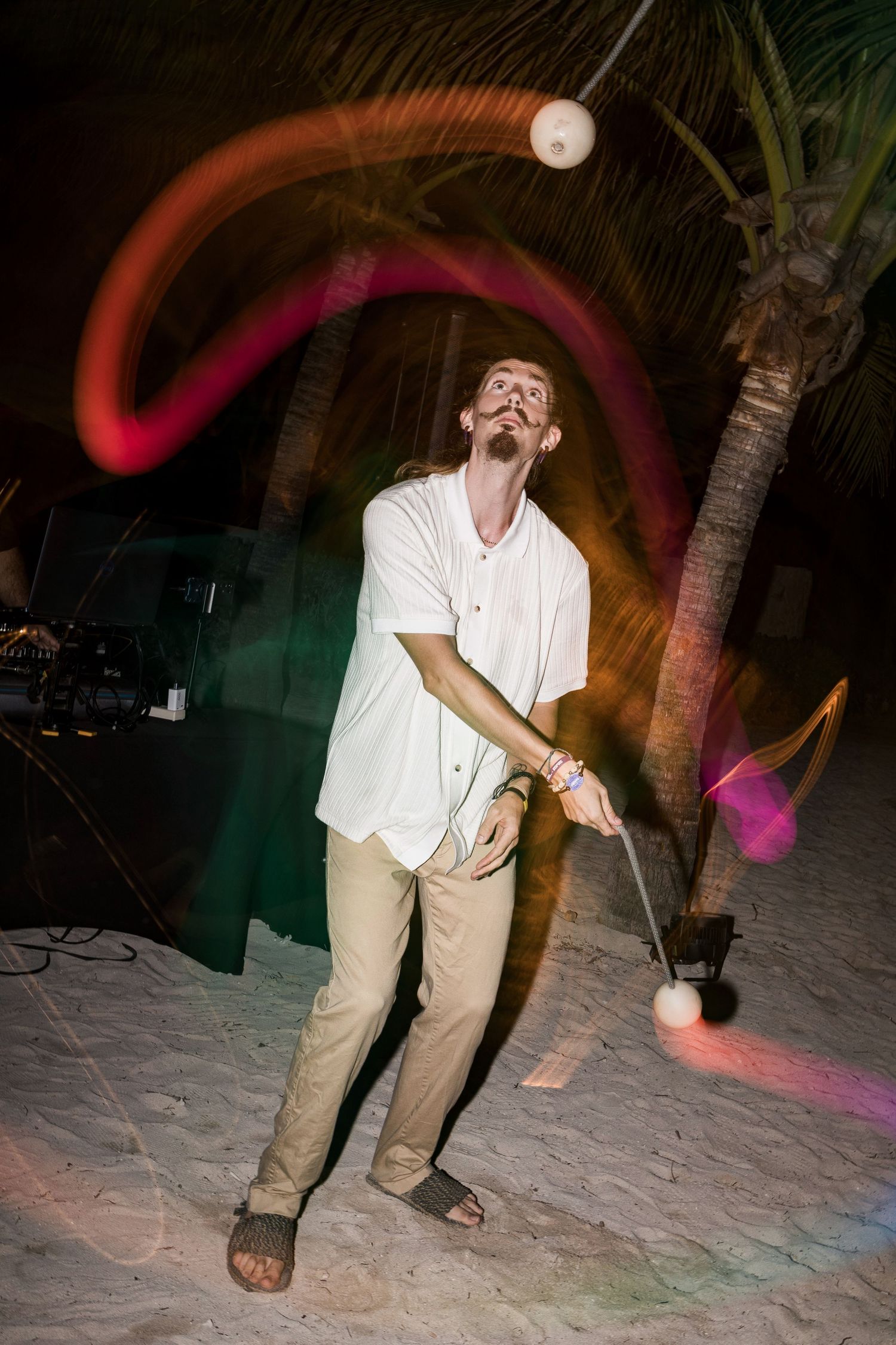 Man juggling glowing balls on a sandy beach at night with colorful light trails and palm trees in background.