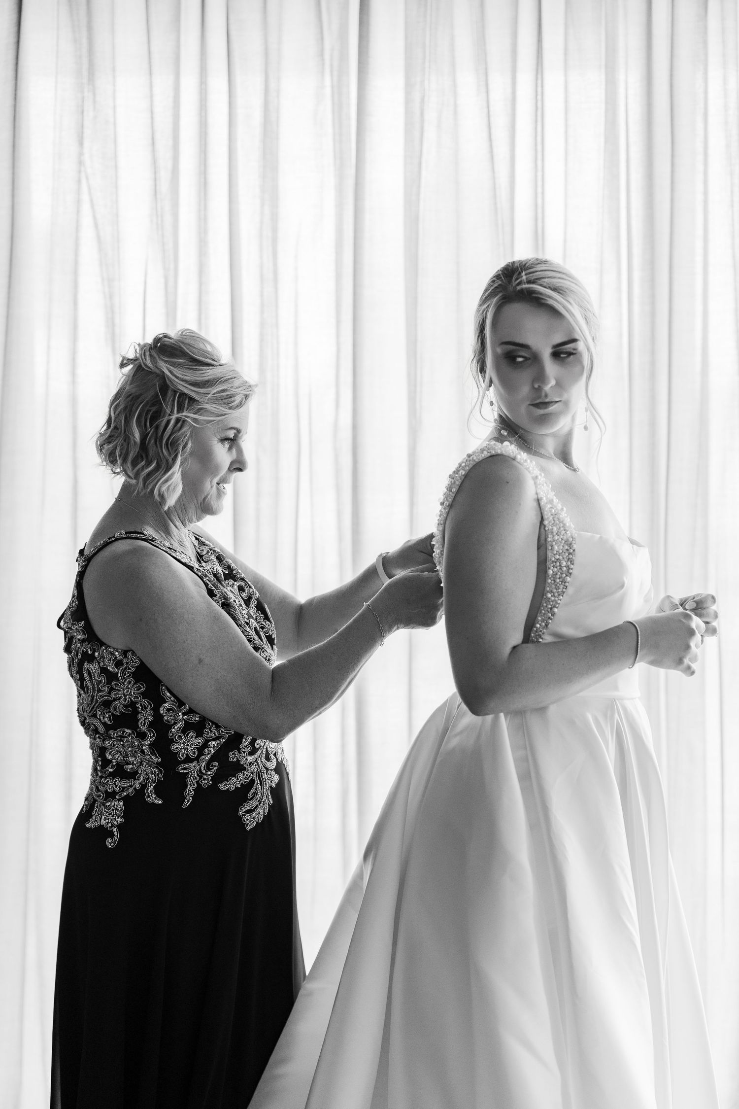 Mother helps bride fasten wedding gown in black and white photo with soft curtain backdrop.