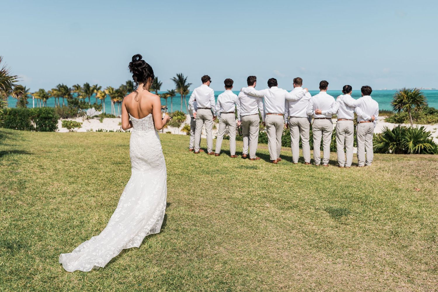 Bride in lace wedding gown stands facing groomsmen in white suits on tropical beach lawn with turquoise ocean backdrop.