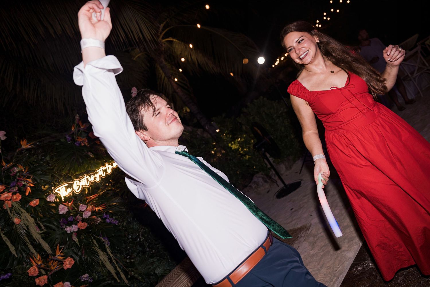 Couple dancing joyfully at outdoor wedding reception, man in white shirt spinning, woman in elegant red gown laughing.