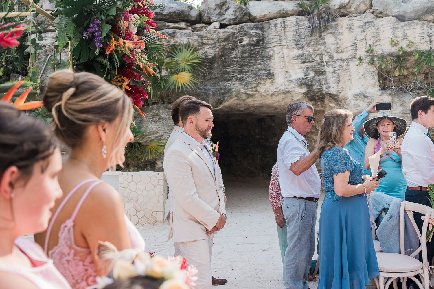 Groom in beige suit stands at outdoor wedding ceremony near cave with tropical floral arch and guests.