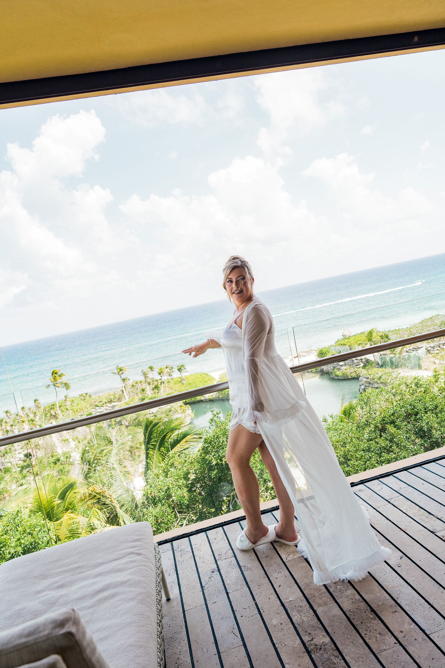 Hotel Xcaret México Wedding  - Bride in white wedding robe standing on tropical resort balcony with ocean view and lush greenery.