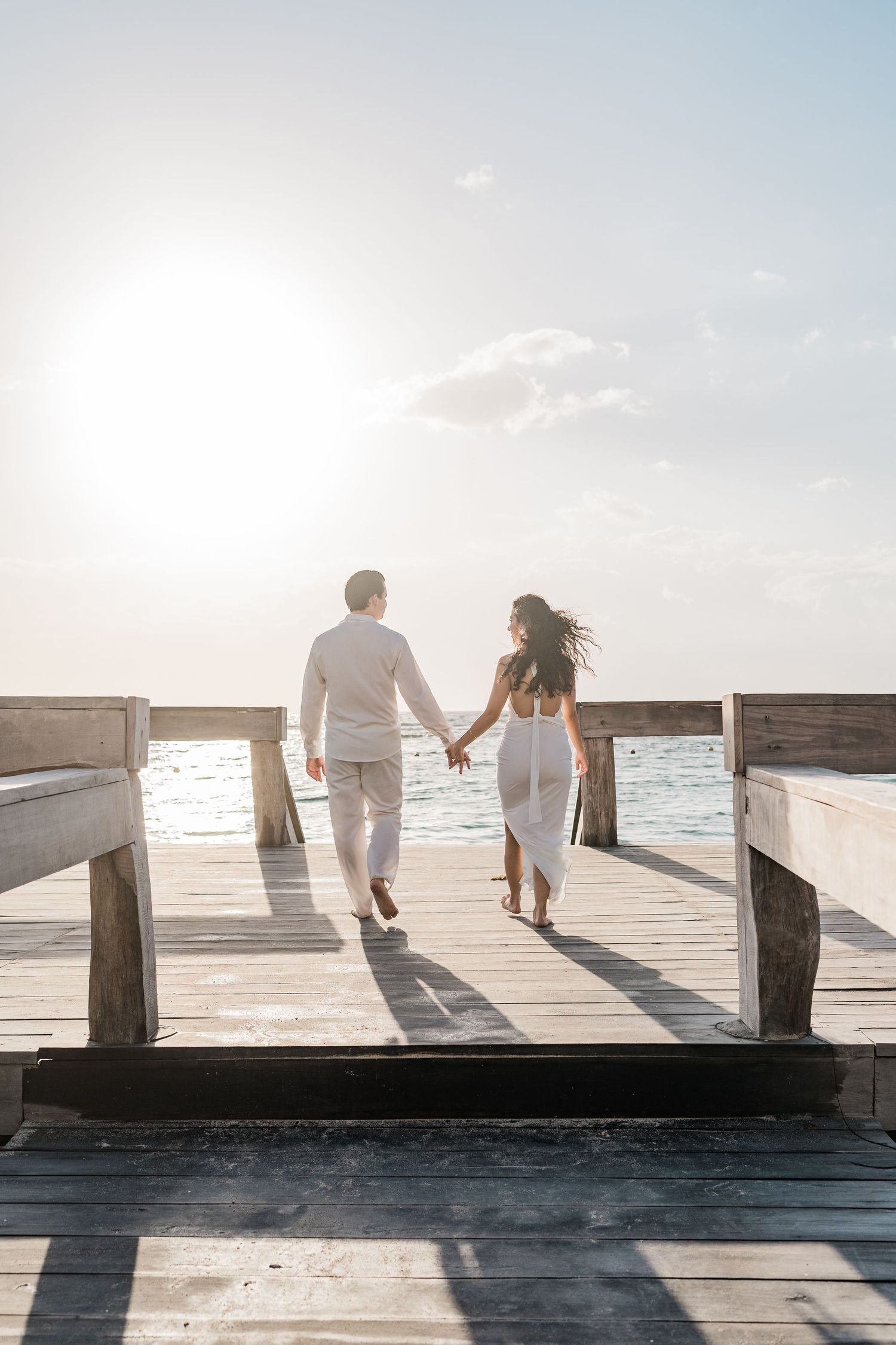 Couple in white outfits holding hands walking on a wooden pier toward the ocean at sunset.