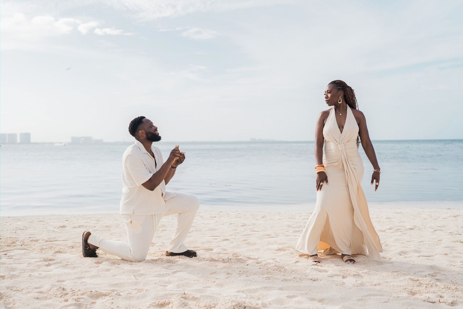 A man proposes on a sandy beach to a woman in a flowing cream dress, with calm ocean waters in the background.