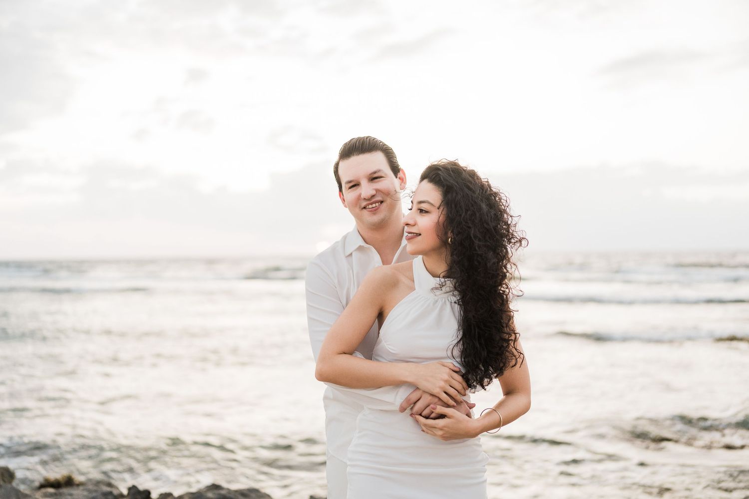Couple embracing on a rocky beach at sunset, wearing white outfits with ocean waves in the background.