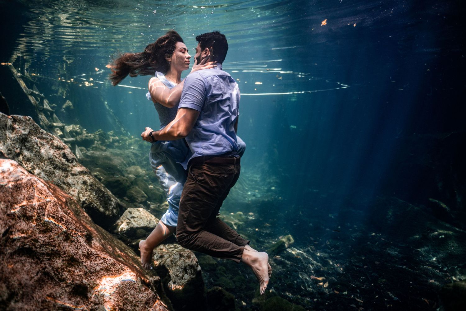 Couple sharing a romantic underwater photoshoot embrace in a crystal-clear cenote, surrounded by rocks and shimmering light - Luis Tovilla