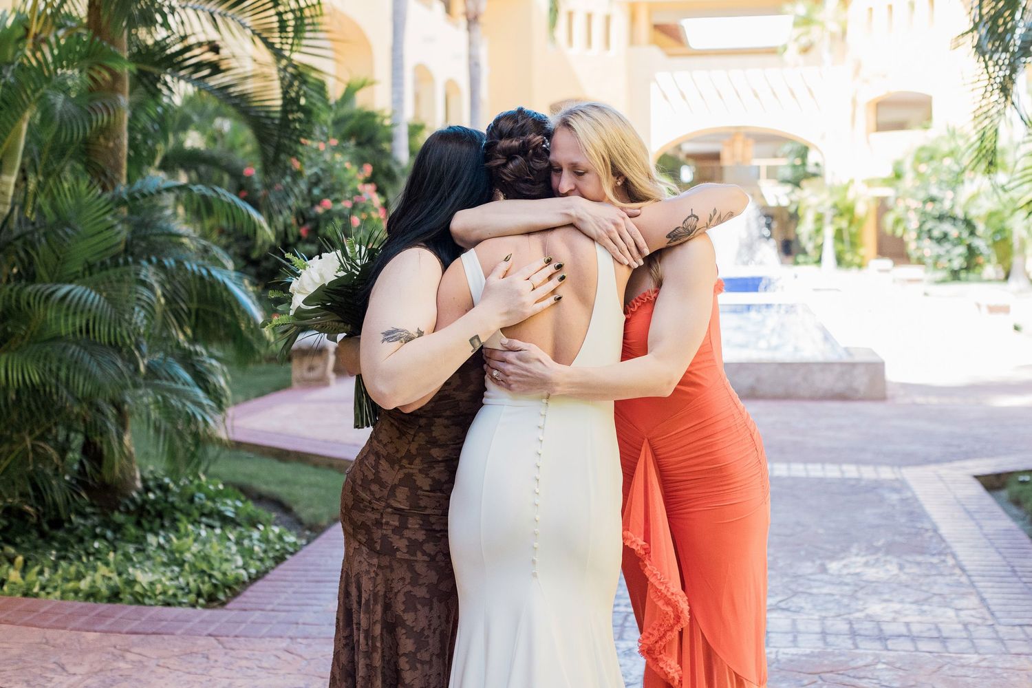 Three women in formal dresses share a warm group hug outdoors at a tropical resort venue.