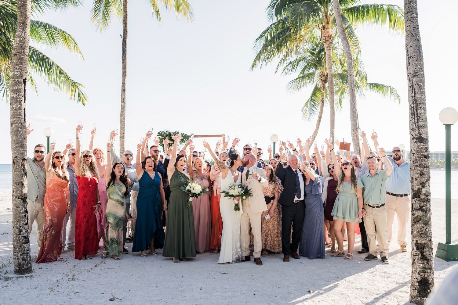 Wedding party celebrates on a tropical beach with palm trees, bridesmaids in colorful gowns, guests cheering with raised arms.