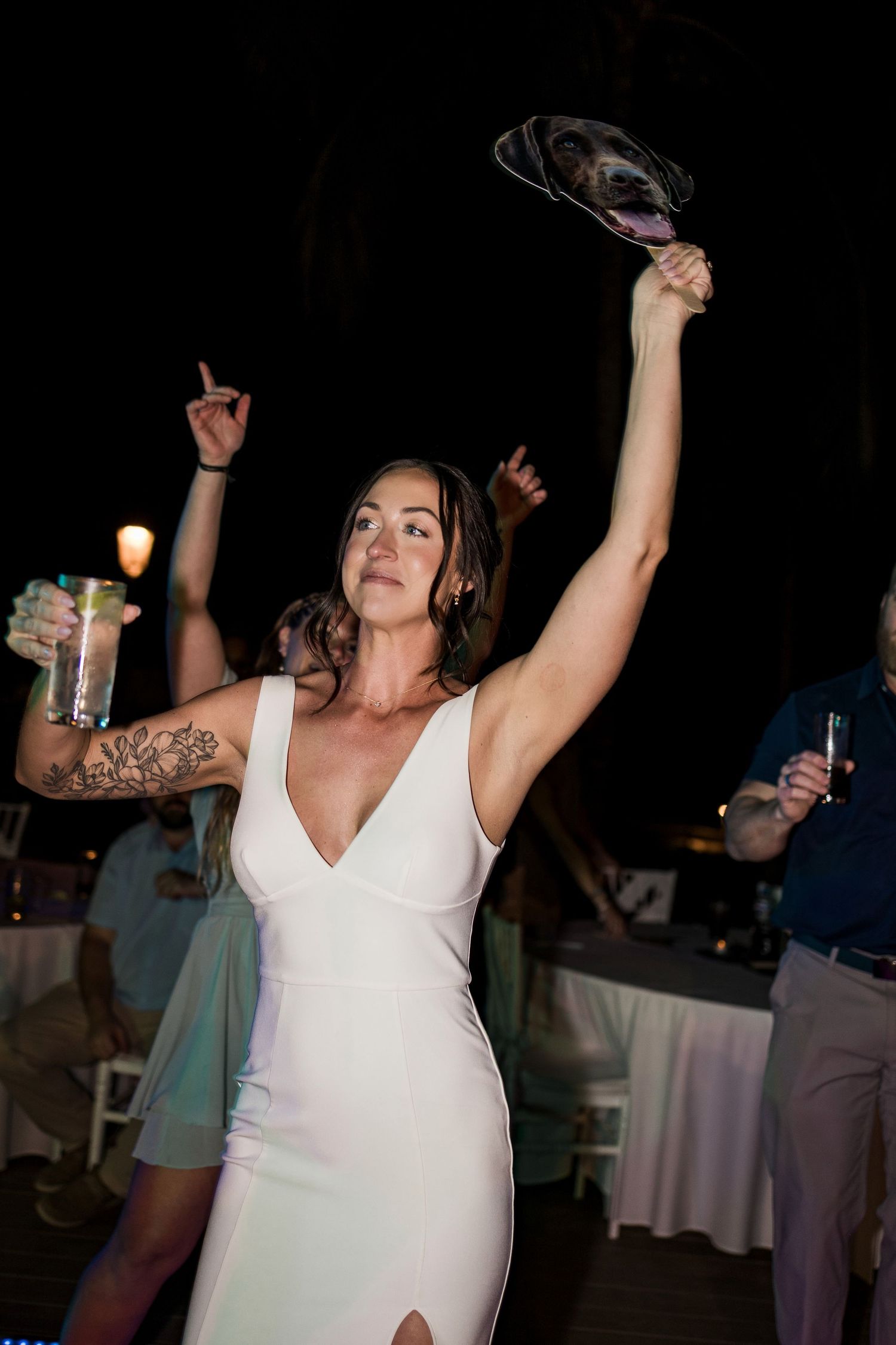 Bride in white dress dancing at wedding reception, holding dog mask above her head while celebrating at night.