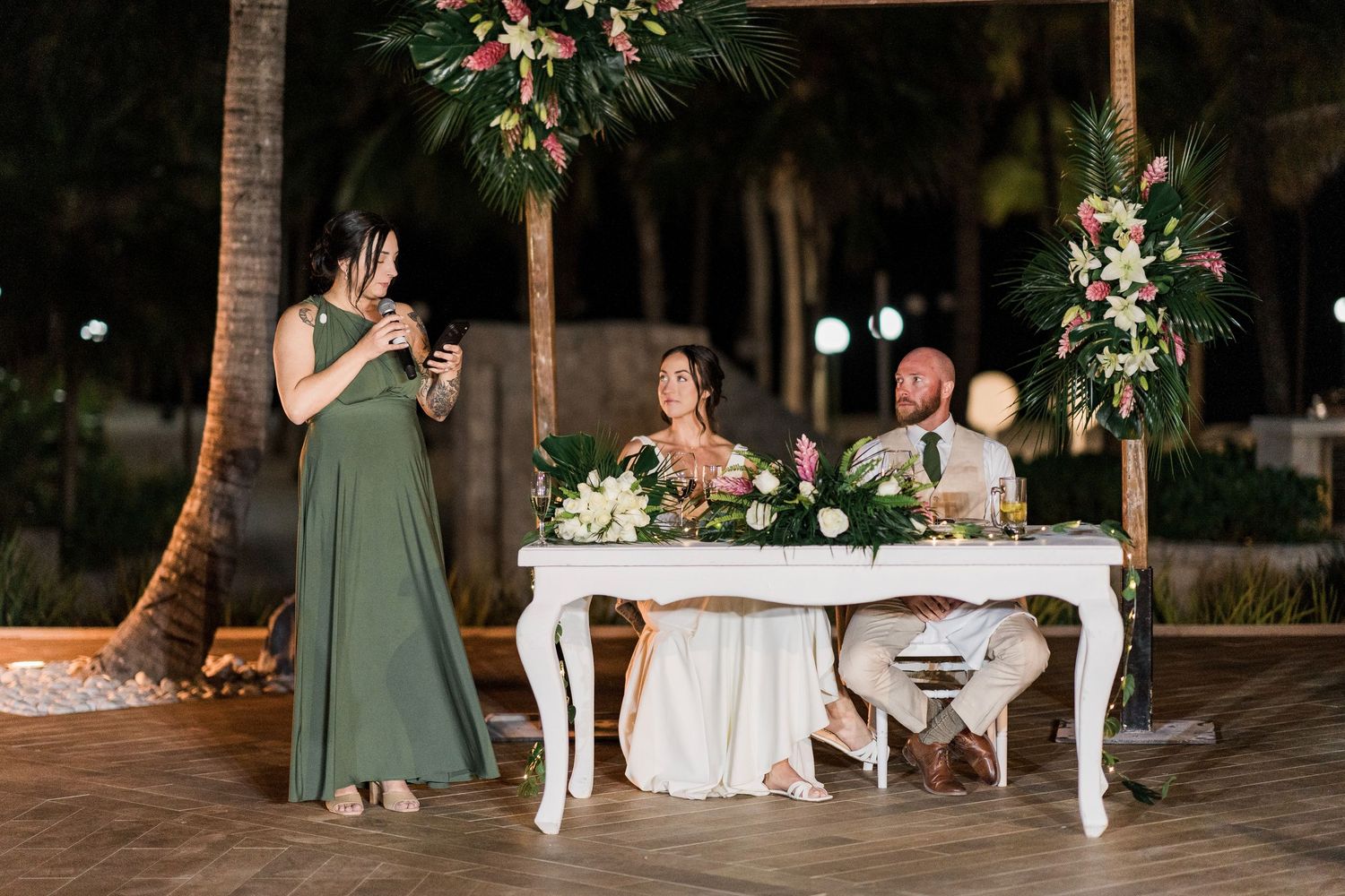 Bridesmaid in green gown gives toast at outdoor tropical wedding reception while couple sits at elegant white sweetheart table.