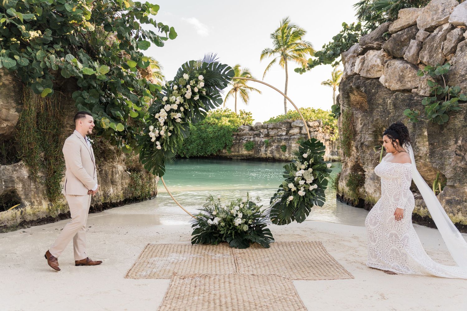 Xcaret Arte Wedding - Couple exchanging vows at a stunning tropical outdoor wedding ceremony by a turquoise lagoon with floral arch decor.