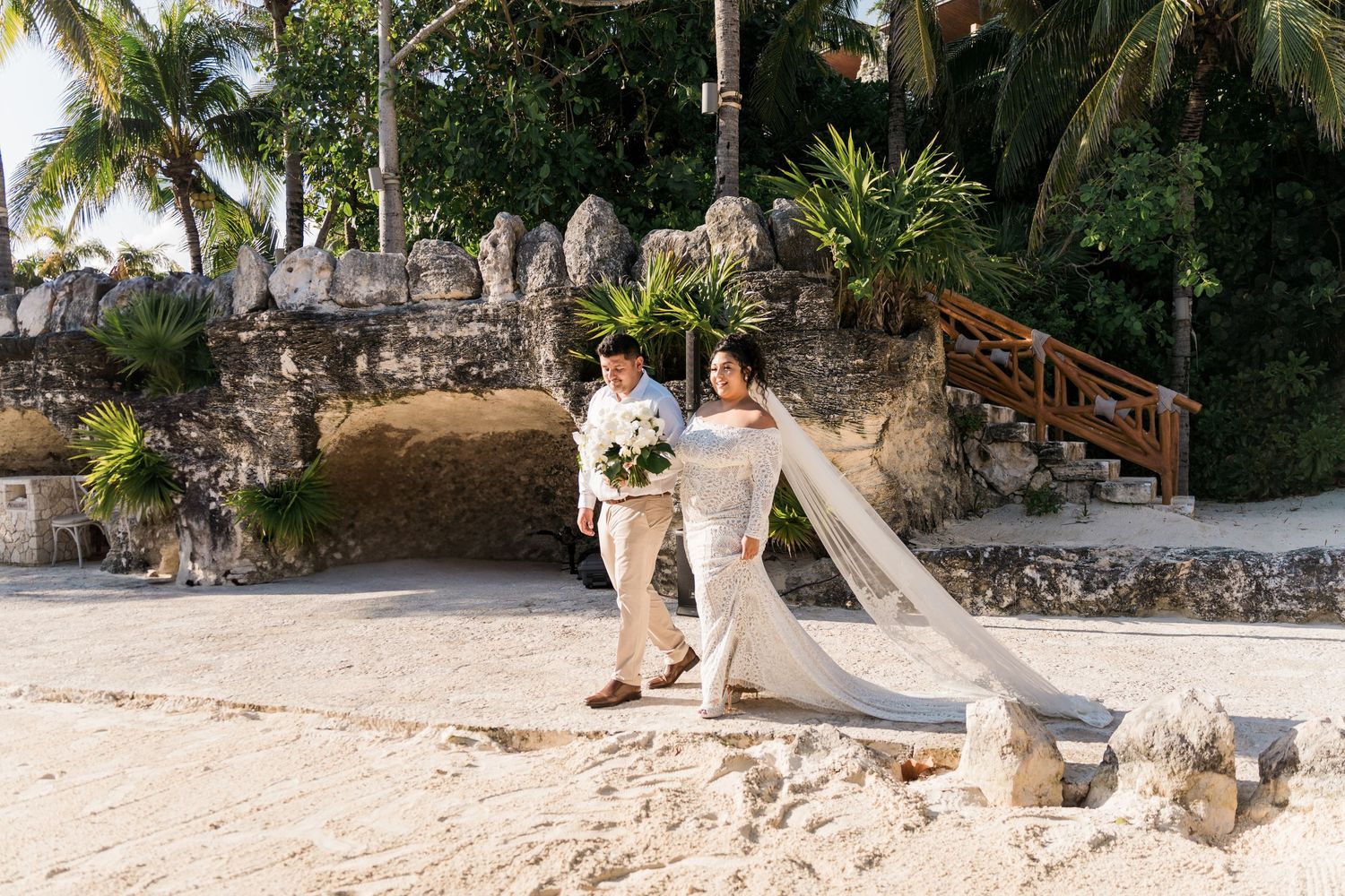 Xcaret Arte Wedding - Bride and groom walk hand-in-hand on white sand beach with tropical greenery and stone ruins backdrop.