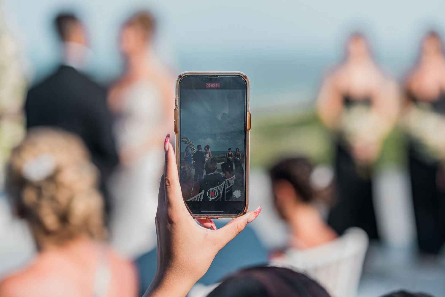 Wedding guest capturing ceremony on smartphone with bride, groom, and wedding party blurred in background.