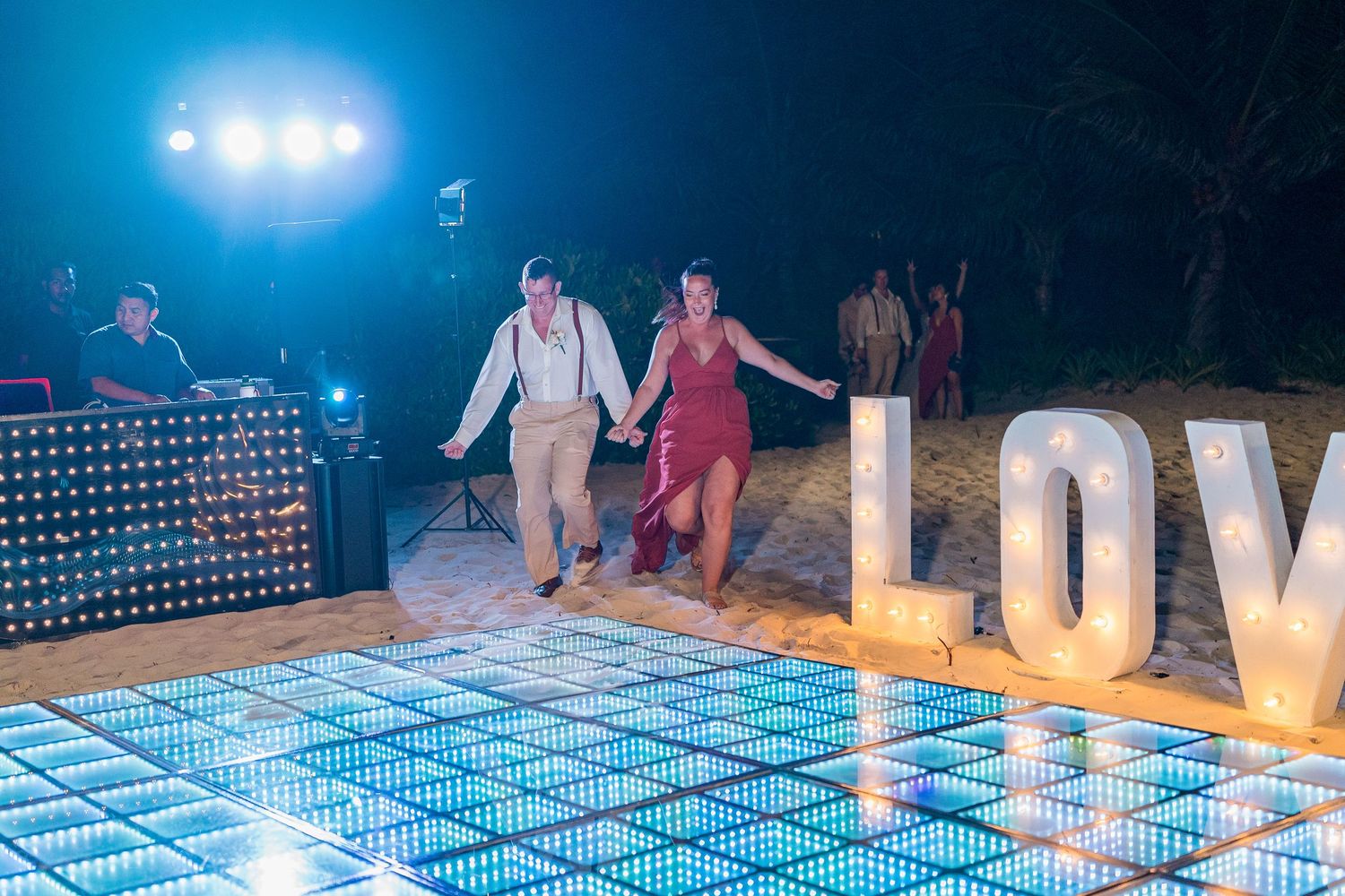 Bride and groom walking hand in hand onto a glowing LED dance floor at their beach wedding reception.