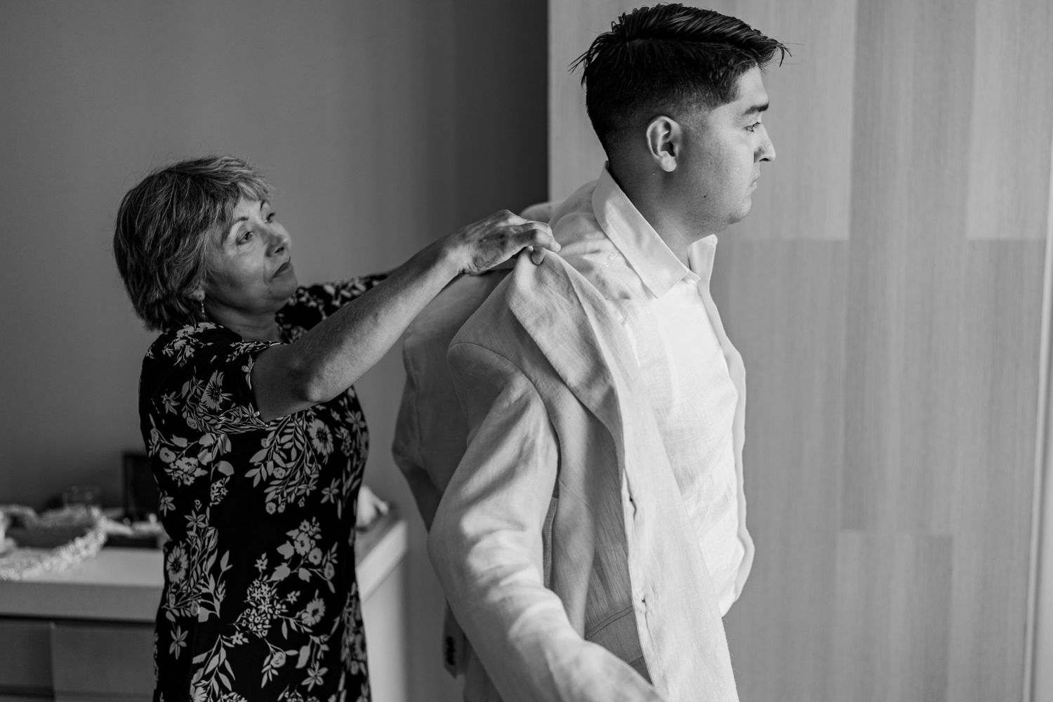 Mother helps groom adjust suit jacket before wedding ceremony in black and white.