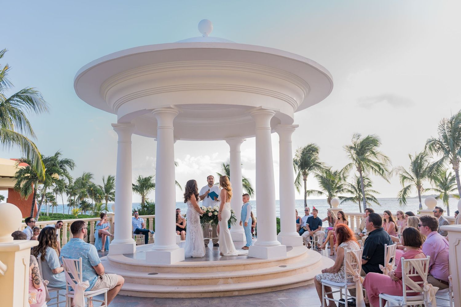 Two brides standing at a white gazebo altar during a tropical beachside wedding ceremony.