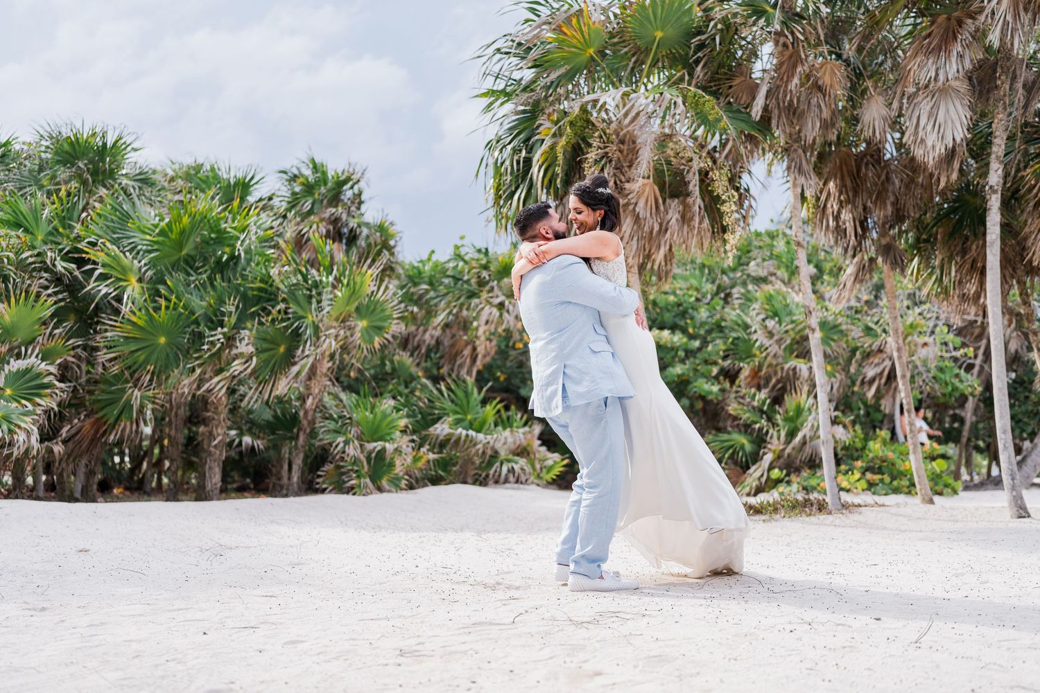 Groom in light blue suit lifts bride in romantic embrace on white sand beach surrounded by palm trees.