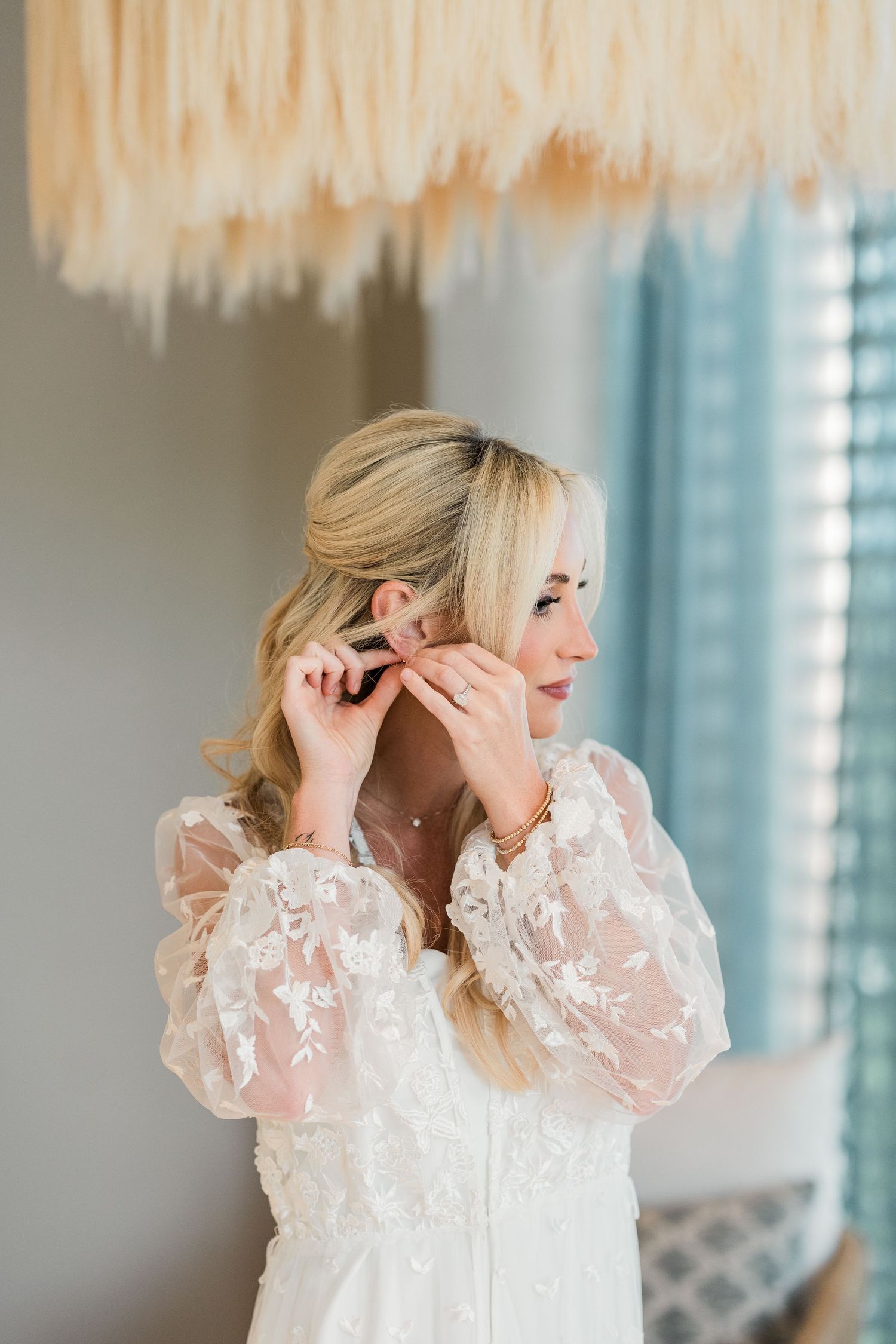 Blonde bride in floral lace robe putting on earrings while getting ready for her wedding day.