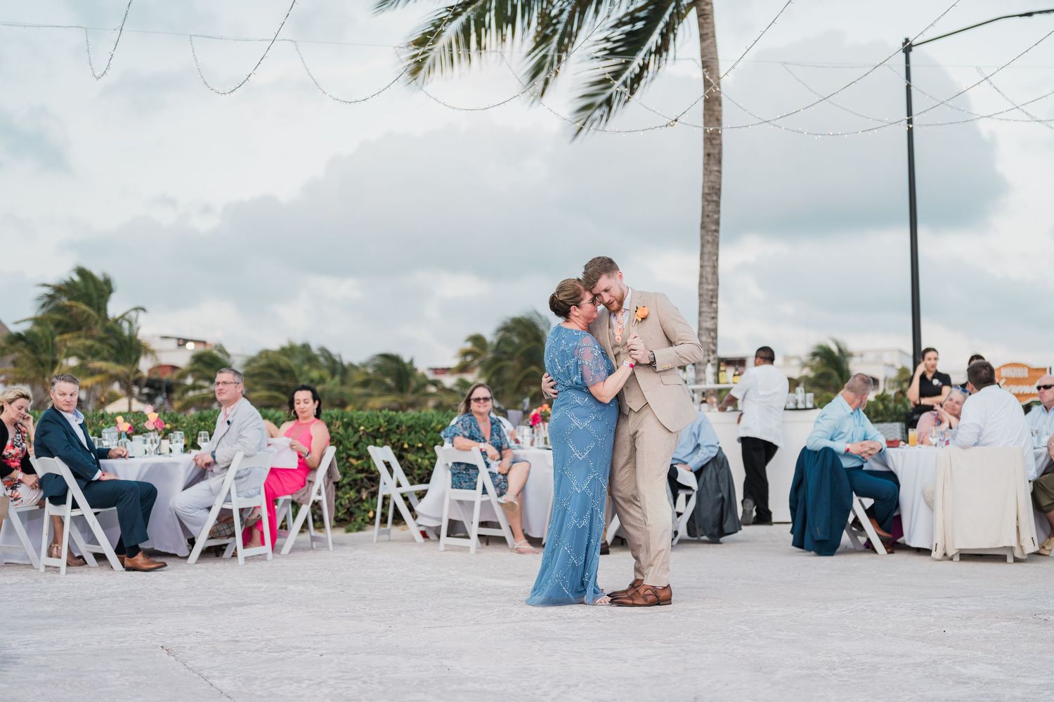Groom shares a tender slow dance with a guest in a blue dress at an outdoor tropical wedding reception.