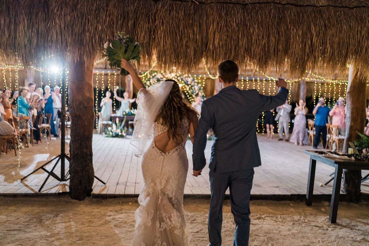 Bride and groom making grand entrance into outdoor palapa wedding reception with cheering guests.