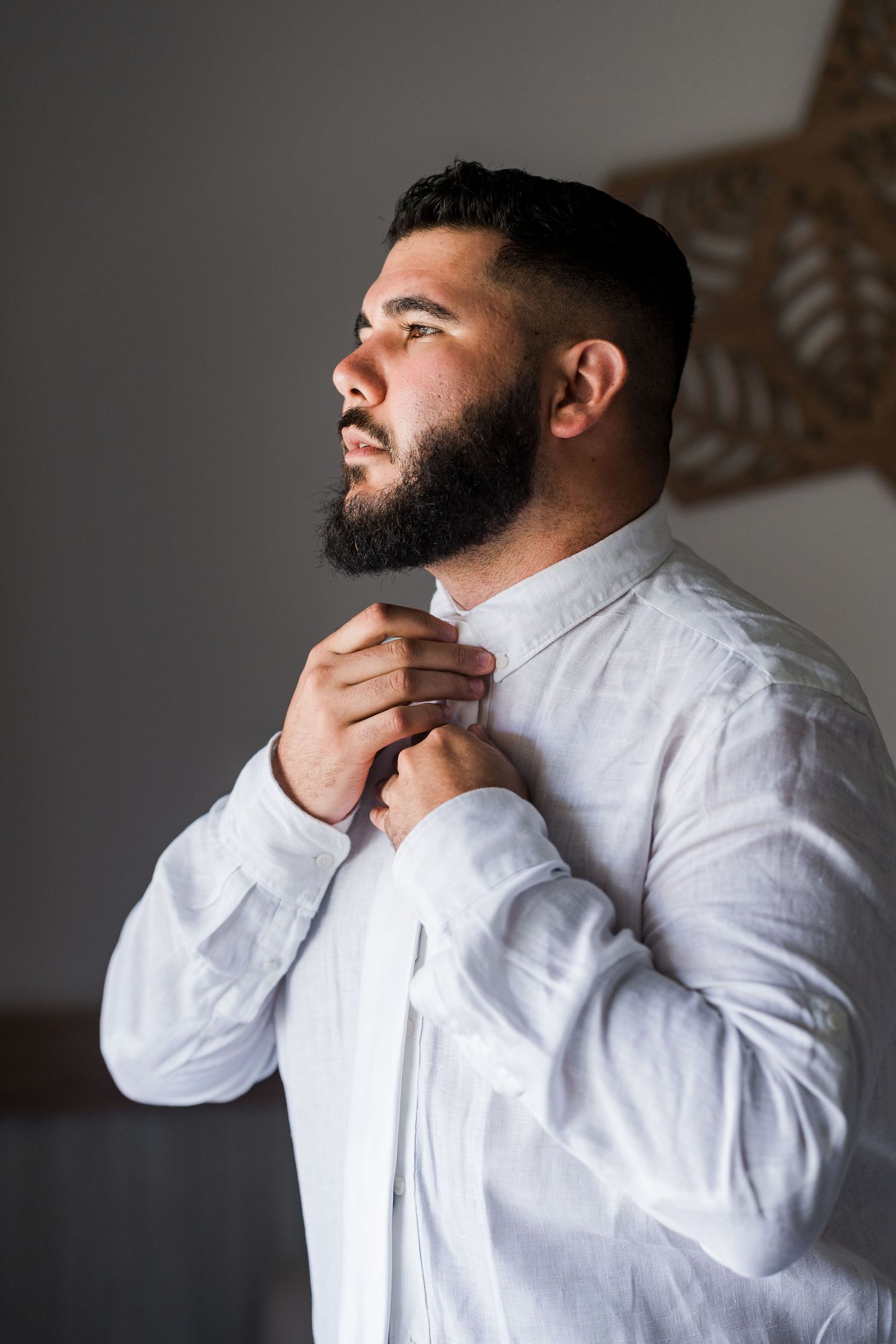 Bearded groom in white linen shirt adjusting his collar while getting ready for his wedding day.